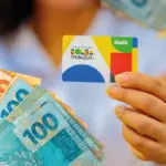 Mãos segurando cartão do Bolsa Família com logotipo colorido e notas de R$ 20 e R$ 100 em reais